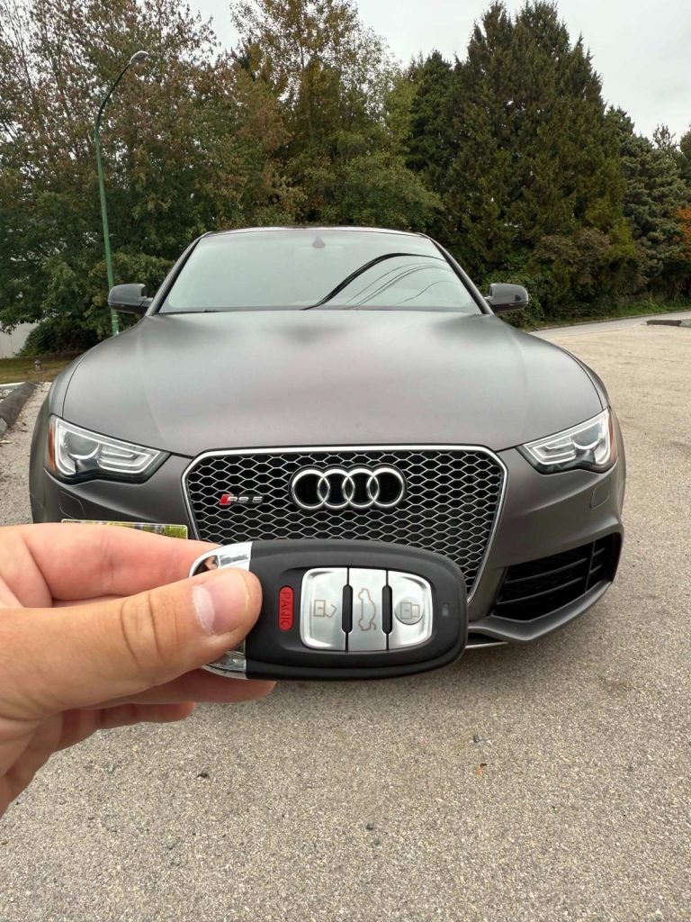 Hand holding an Audi smart key fob in front of a gray Audi coupe