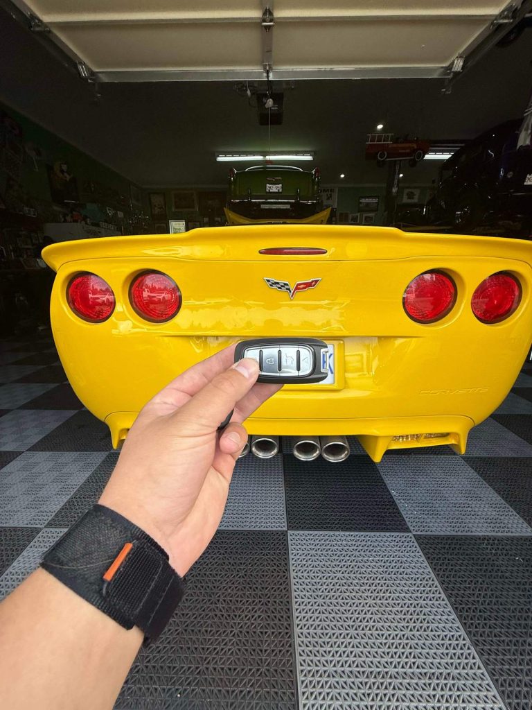Hand holding a key fob at the rear of a yellow Chevrolet Corvette in a garage