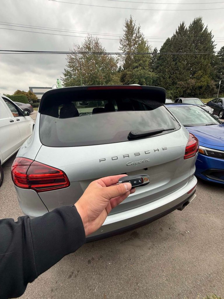Hand holding a key fob behind a silver Porsche Cayenne SUV in a parking lot