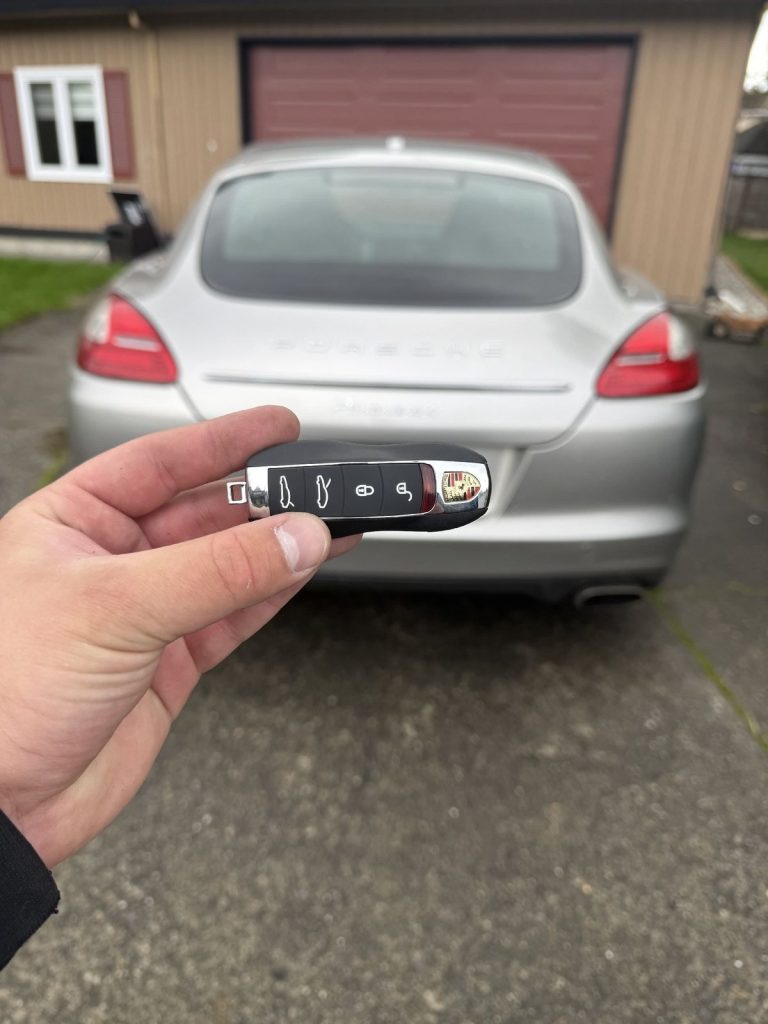 Hand holding a Porsche key fob behind a silver Porsche Panamera parked outside a garage
