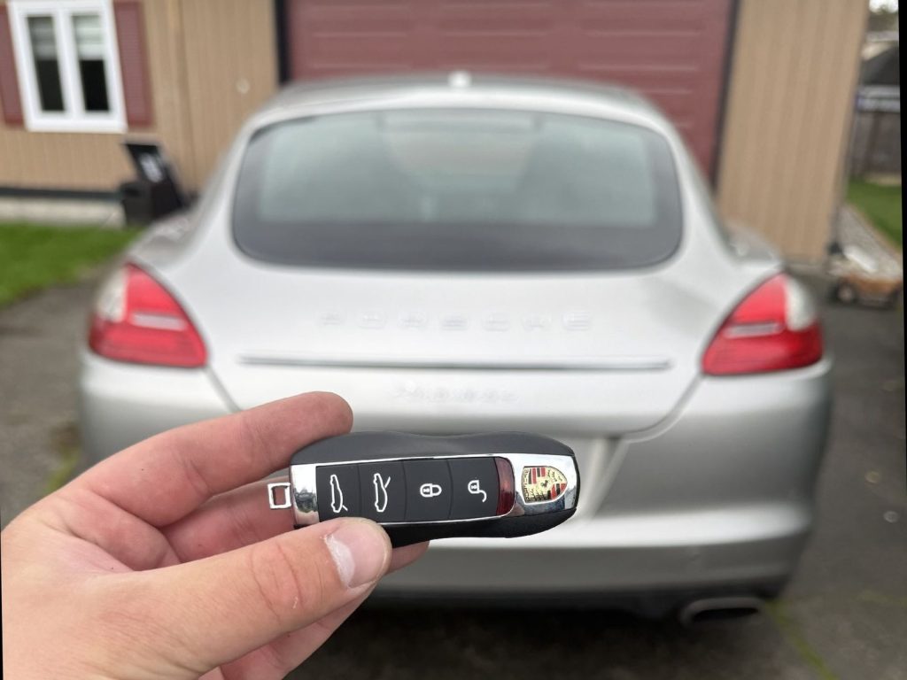 Hand holding a Porsche smart key fob in front of the rear of a silver Porsche Panamera