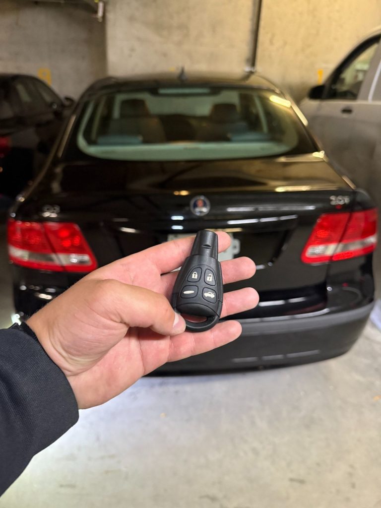 Hand holding a key fob behind a black Saab sedan parked in a garage